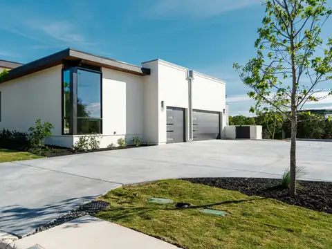 Wide broom-finish concrete driveway leading to a modern white house with a dark garage door and landscaping