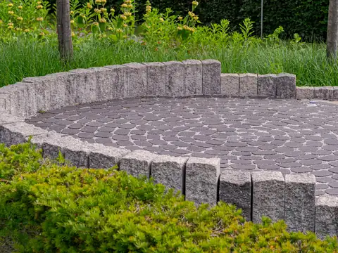 Circular paver patio bordered by granite edging stones in a landscaped garden with shrubs and tall grass